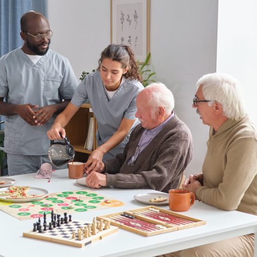 Nurse and doctor pouring hot tea for senior people while they sitting at the table and playing board games in nursing home