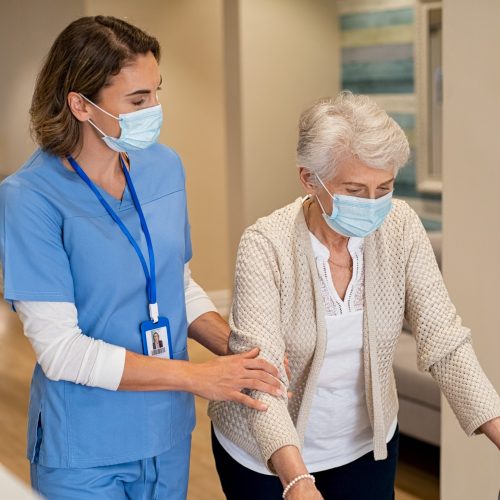 Smiling nurse with face mask helping senior woman to walk around the nursing home with walker. Young lovely nurse helping old woman with surgical mask for safety against covid-19 using a walking frame in hospital corridor. Friendly caregiver and elderly patient walking in hallway wearing protective face mask during covid pandemic.