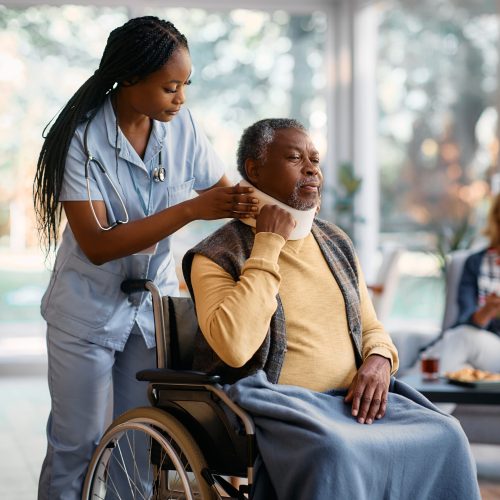 African American nurse putting cervical collar on senior man's neck at nursing home.